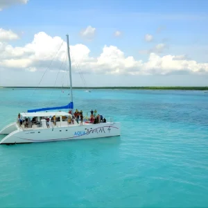 Un catamarán blanco llamado Aqua Dream 4 navegando por las aguas turquesas y tranquilas de Cozumel, México, con varios pasajeros a bordo disfrutando del recorrido bajo un cielo parcialmente nublado.